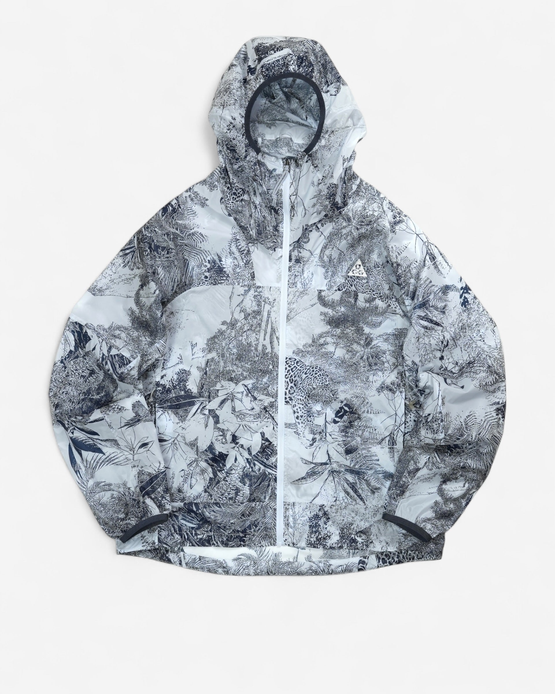  Світло-сіра вітровка Nike ACG Cinder Cone Printed Windproof Jacket з капюшоном, контрастною графікою джунглів та білим трикутним логотипом на грудях.