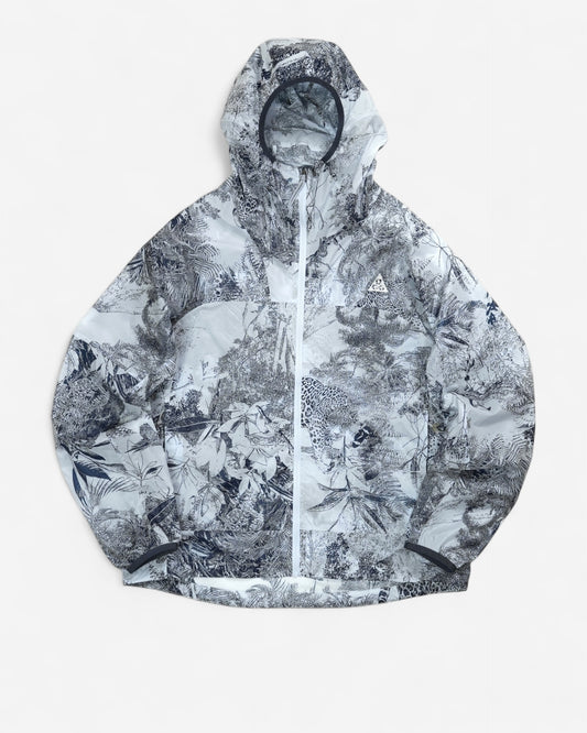  Світло-сіра вітровка Nike ACG Cinder Cone Printed Windproof Jacket з капюшоном, контрастною графікою джунглів та білим трикутним логотипом на грудях.