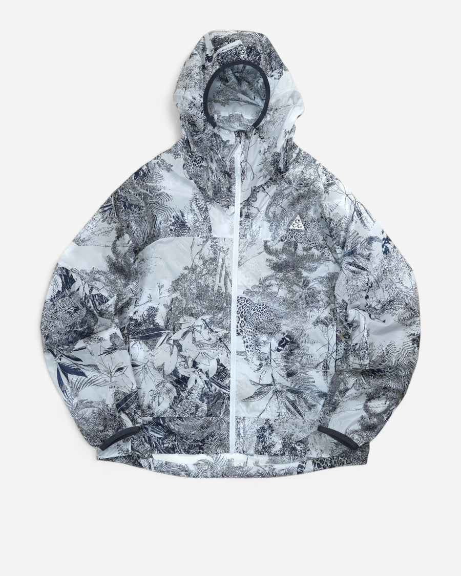  Світло-сіра вітровка Nike ACG Cinder Cone Printed Windproof Jacket з капюшоном, контрастною графікою джунглів та білим трикутним логотипом на грудях.