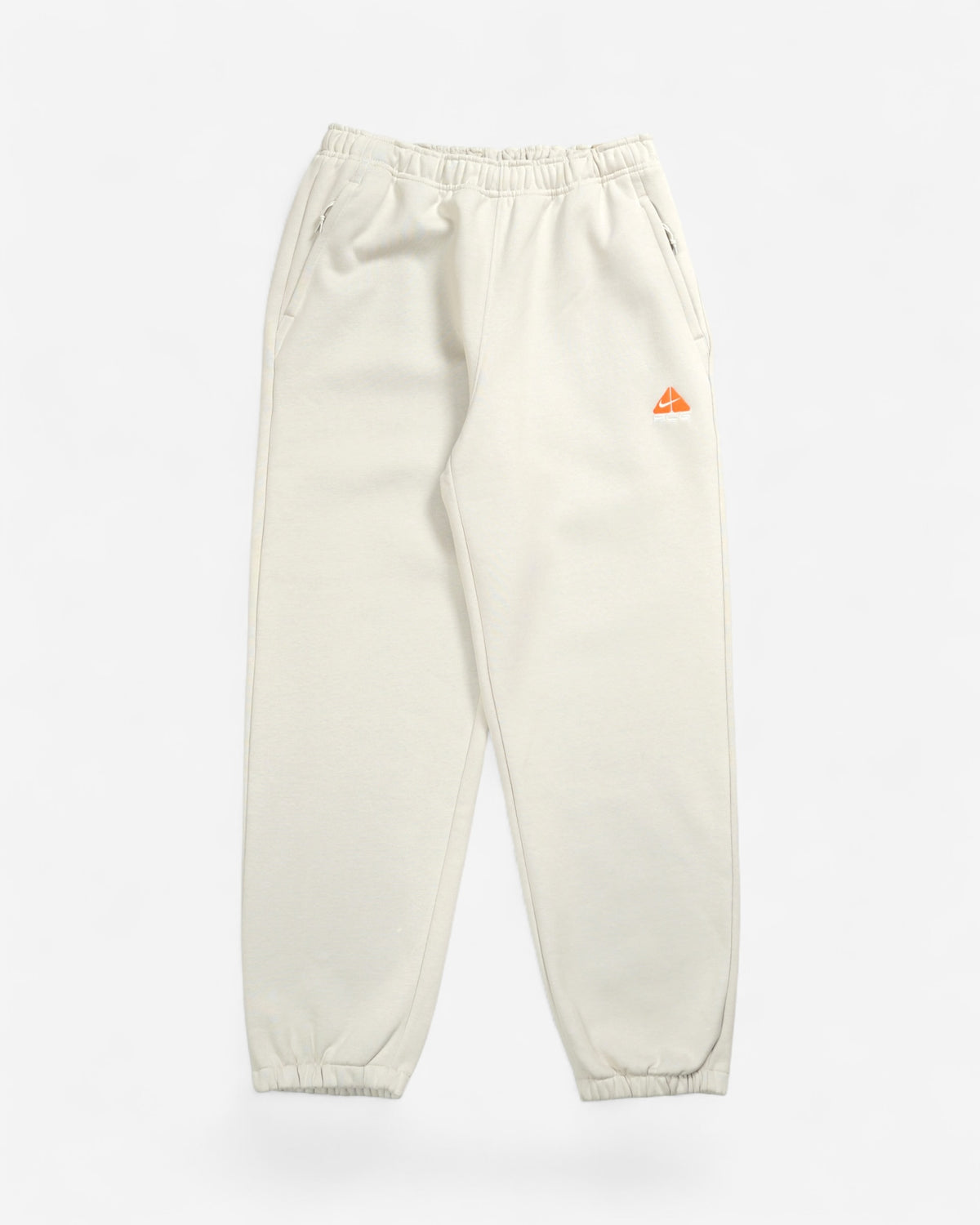 Світло-бежеві спортивні штани Nike ACG Tuff Fleece з еластичним поясом, манжетами та яскравим помаранчевим вишитим трикутним логотипом на лівому стегні.