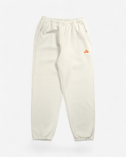 Світло-бежеві спортивні штани Nike ACG Tuff Fleece з еластичним поясом, манжетами та яскравим помаранчевим вишитим трикутним логотипом на лівому стегні.