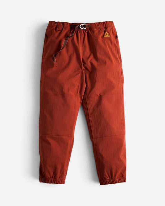 Теракотово-руді штани Nike ACG Trail Pants з еластичним поясом, вбудованим темним ременем та фірмовим трикутним логотипом на лівому стегні.