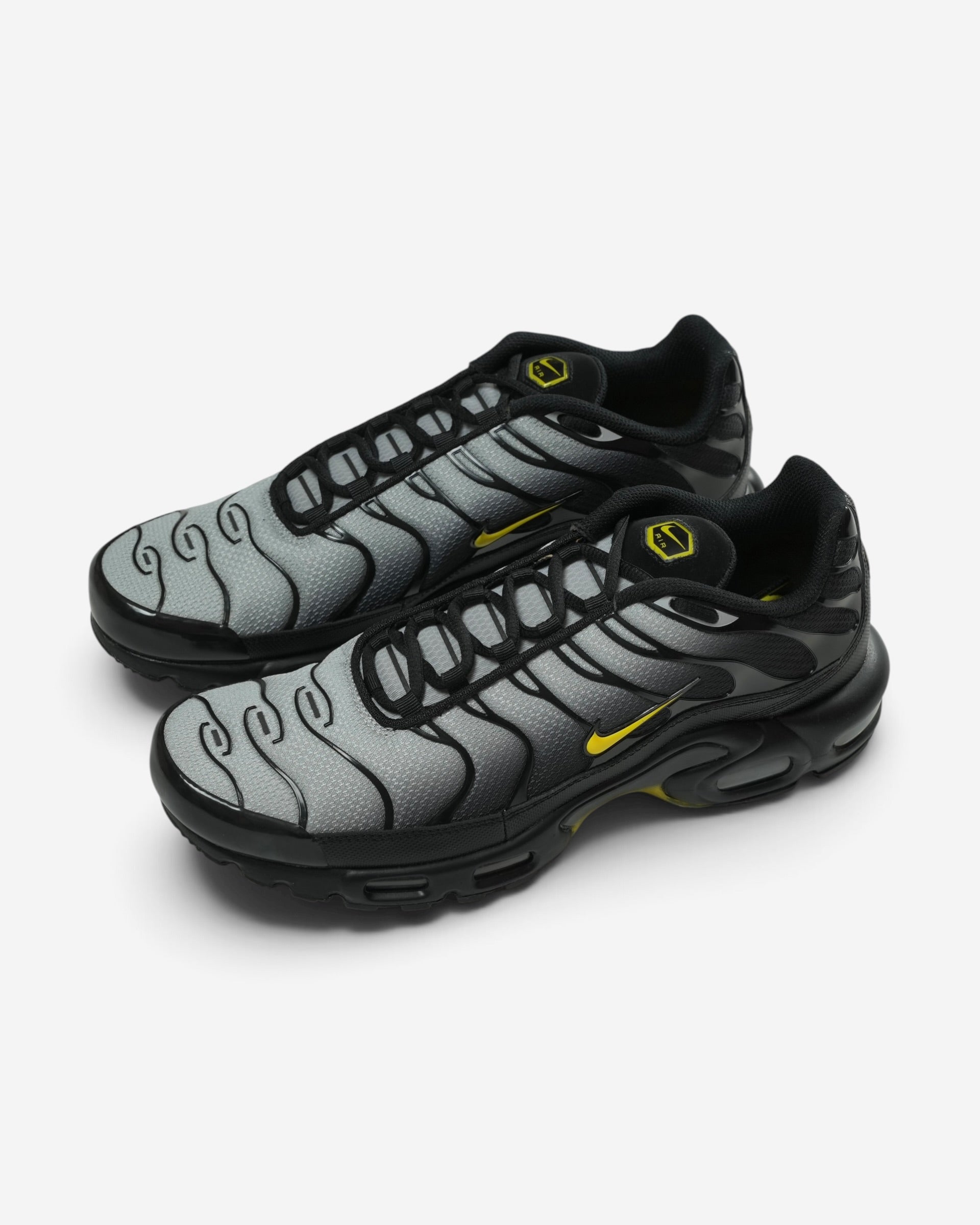 Чорно-сірі кросівки Nike Air Max Plus із плавним градієнтним переходом, тонкими гнучкими лініями каркаса та яскраво-жовтими фірмовими логотипами.