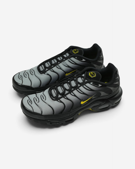 Чорно-сірі кросівки Nike Air Max Plus із плавним градієнтним переходом, тонкими гнучкими лініями каркаса та яскраво-жовтими фірмовими логотипами.