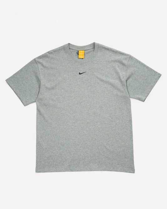 Меланжева сіра футболка вільного оверсайз крою Nike x NOCTA з маленьким чорним свушем по центру грудей та помаранчевою біркою на горловині.