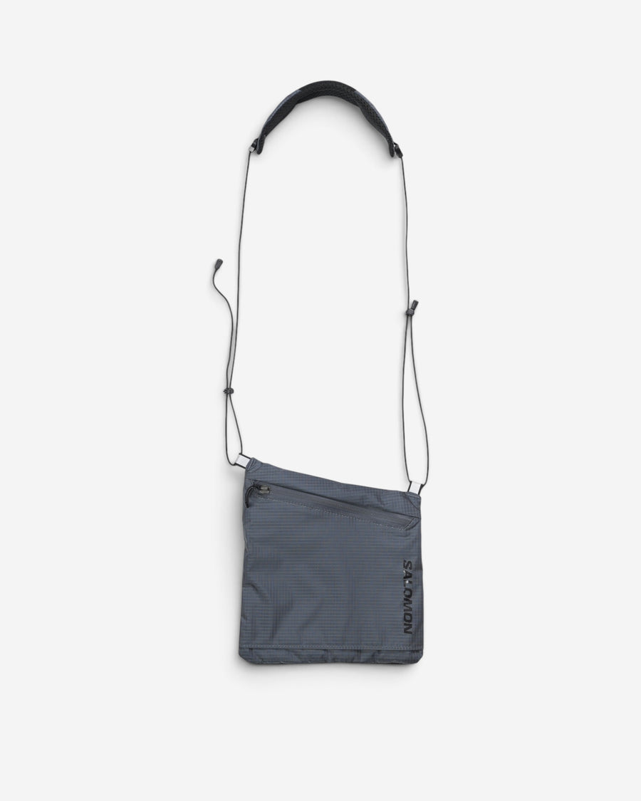 Сіра невелика сумка через плече Salomon ACS Pouch 2 із тонким шнуром-ременем, плечовою накладкою та чорним вертикальним логотипом бренду.